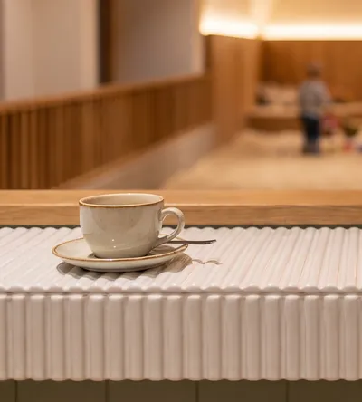 Ein Cappuccino auf dem gerillten Tresen, dahinter warm beleuchtet die Sandhalle.